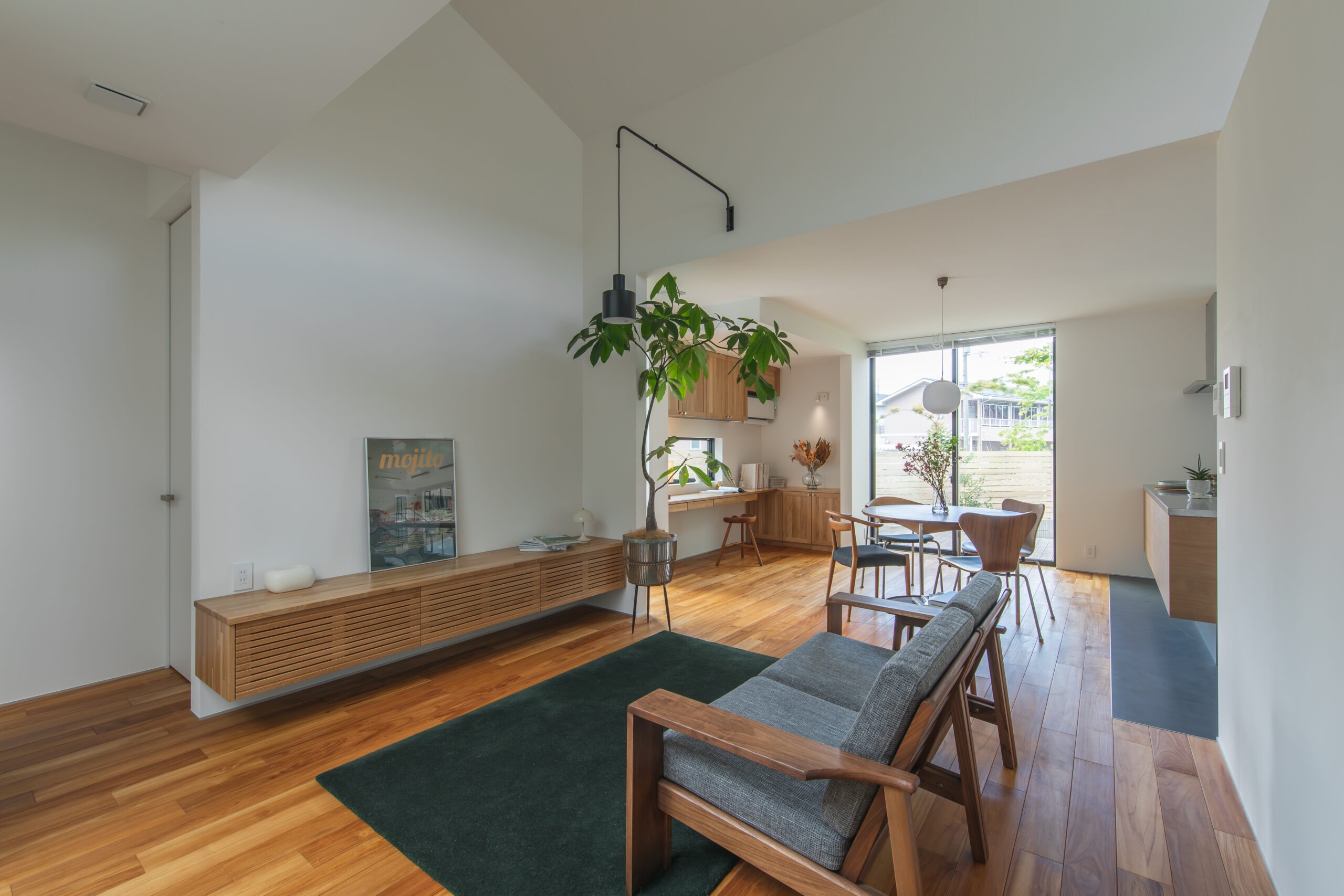 Open-concept living area with a wooden sideboard, tall plant, and a round dining table near a large window.