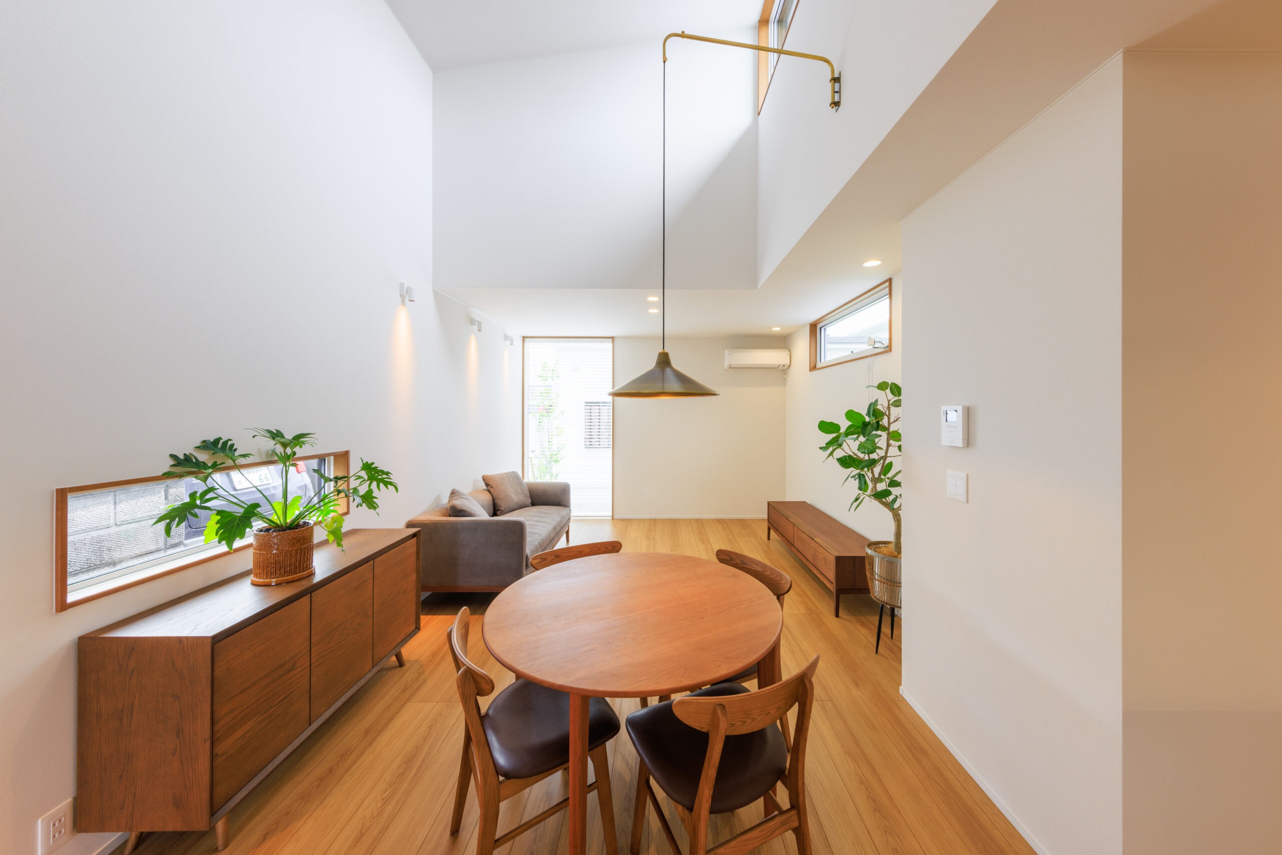 Bright open-plan living/dining area with round wooden table and chairs, low wooden credenza, and potted plants along white walls.